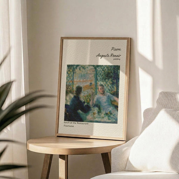 May include: Framed art print of Renoir's impressionist painting showing two figures dining at a restaurant with lattice work and foliage. Print has cream background with black text identifying the artist and artwork title. Displayed on round wooden side table next to beige upholstered chair, with plant leaves visible and natural sunlight casting shadows on white wall behind.