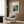 May include: A framed Renoir poster print hanging on wood paneled wall above a beige armchair with geometric pillow. The vintage-style art poster shows an impressionist restaurant scene with people dining outdoors in soft blues and greens. Modern brass floor lamp and black side table with open book complete the neutral-toned reading nook setting.