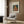 May include: Framed Renoir poster print showing red-haired girl in coral dress sewing, mounted on wood panel wall above beige upholstered armchair with geometric pillow. Modern brass floor lamp and black side table with open book complete the neutral-toned living room scene with wood flooring and white curtains.
