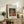 May include: Framed art print of young girl with red hair sewing, displayed on rustic wood sideboard beside fern plant, ceramic vase, and stacked books in neutral-toned living room with beige armchair and throw pillows