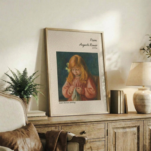 May include: Framed art print of young girl with red hair sewing, displayed on rustic wood sideboard beside fern plant, ceramic vase, and stacked books in neutral-toned living room with beige armchair and throw pillows