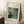 May include: Framed poster print of Renoir's impressionist painting showing two figures dining at a restaurant terrace with latticed windows. The artwork features soft blues and greens with dappled light effects. Light wood frame with white matting, leaning against white wall corner on textured cream rug. Coffee cup on wooden block visible nearby, creating cozy home interior setting with natural window light casting shadows.