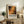 May include: Abstract art poster with warm autumn colors - oranges, yellows, and dark greens in chaotic splattered paint style, displayed in light wood frame on rustic wooden sideboard. Room scene shows cream upholstered chair, potted fern, ceramic vase, stacked books, and soft natural lighting against white walls.