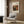 May include: Cream-colored framed art print featuring Renoir's still life painting of pomegranate and figs in warm oranges and greens, displayed on wood-paneled wall above beige upholstered armchair with geometric patterned pillow, brass floor lamp and dark round side table with open book in modern neutral-toned room setting.