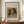 May include: Framed vintage dog portrait print displayed on wooden sideboard next to white ceramic vase and books. The reproduction features a small spaniel-type dog with long ears and collar against muted brown background. Cream matting surrounds the artwork in thin wooden frame. Soft natural lighting illuminates the elegant interior setting with white paneled walls.