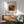 May include: Framed Renoir art print featuring pomegranates and figs in warm orange, green, and brown tones, displayed above a wooden sideboard in a modern living room with mint green armchair, white table lamp, and natural sunlight creating geometric shadows on white walls.