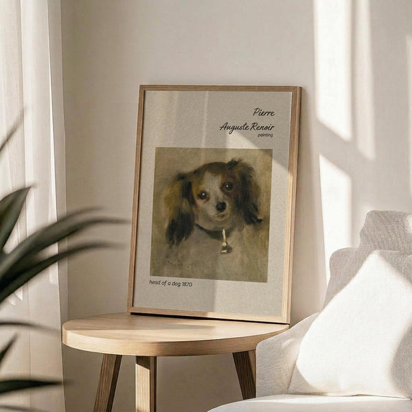 May include: Framed art print featuring Renoir's 1870 painting of a small dog with dark and light brown fur and a collar, displayed on a round wooden side table next to a beige upholstered chair. Soft natural light creates gentle shadows on the neutral wall behind. A plant is partially visible in the corner, creating a cozy living room setting with warm, minimalist decor.