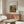 May include: Framed poster of Pierre Auguste Renoir's 1917 Farmhouse painting displayed above a wooden sideboard in a minimalist living room. The colorful impressionist artwork shows a Mediterranean landscape with warm earth tones, green foliage, and architectural elements. Modern furniture includes a cream curved chair, glass oval coffee table, and brass floor lamp with circular mirror detail.