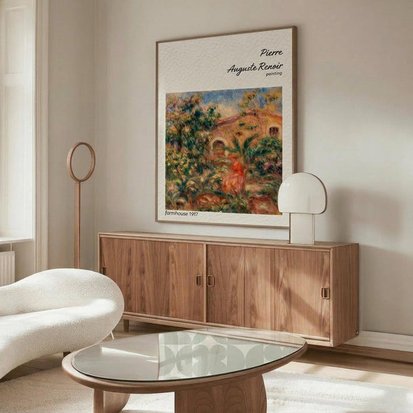 May include: Framed poster of Pierre Auguste Renoir's 1917 Farmhouse painting displayed above a wooden sideboard in a minimalist living room. The colorful impressionist artwork shows a Mediterranean landscape with warm earth tones, green foliage, and architectural elements. Modern furniture includes a cream curved chair, glass oval coffee table, and brass floor lamp with circular mirror detail.