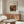 May include: Renoir still life poster showing pomegranates and figs in warm reds, oranges and greens, framed and displayed above a walnut wood credenza in a minimalist living room. White curved chair and glass-top oval coffee table visible. Natural lighting creates soft shadows on cream walls.