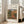 May include: Framed art print of Pierre-Auguste Renoir's impressionist landscape painting showing figures in a lush garden setting with warm earth tones, greens, and blue sky. Light wood frame with white matting, leaning against beige wall next to cream-colored armchair in bright, naturally-lit living room with white trim and hardwood floors.