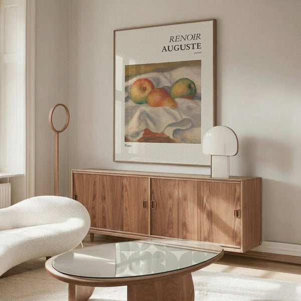 May include: Framed art print of Auguste Renoir's pears painting displayed on wooden credenza in modern living room. Print shows colorful painted pears on white cloth with cream mat and thin frame. Room features curved white chair, glass oval coffee table, white table lamp, and wooden floor mirror, all in warm neutral tones with natural lighting.