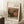 May include: Framed art print leaning against white wall corner on textured cream rug. Light wood frame contains vintage-style still life painting of pomegranate and figs in warm earth tones with green foliage. White coffee cup on wooden block sits nearby. Beige fabric draped in corner. Natural lighting creates soft shadows on white walls.
