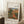 May include: Framed poster print of an impressionist painting showing a figure in red clothing sitting in a lush garden landscape with warm earth tones, greens, and a rustic building in the background. The print is displayed in a light wooden frame, leaning against a white wall next to a beige armchair, with a coffee cup on a wooden side table and white textured rug beneath.