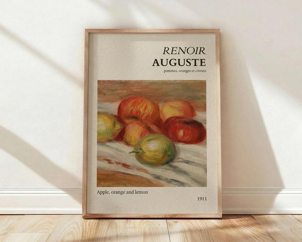 May include: Framed art print leaning against white wall on light wood floor. Cream-colored poster features Auguste Renoir's impressionist still life painting of red apples, yellow orange, and green lemon on draped fabric. French and English text above and below artwork. Natural wood frame with soft window light creating diagonal shadows across the scene.