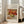 May include: Framed art print leaning against beige sofa in bright living room with large windows. Print shows Renoir's impressionist still life painting of red apples, yellow citrus fruits on white cloth surface. White frame with cream matting, museum-style text above and below artwork. Soft natural lighting creates gentle shadows on hardwood floor.