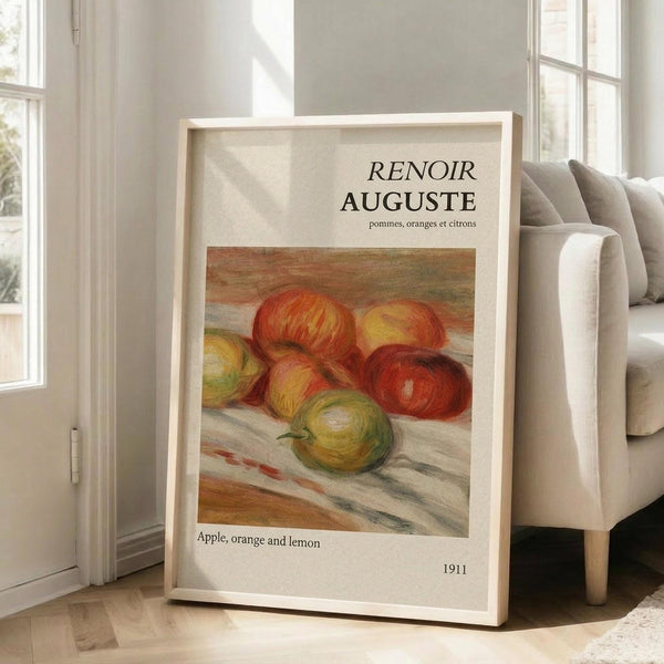 May include: Framed art print leaning against beige sofa in bright living room with large windows. Print shows Renoir's impressionist still life painting of red apples, yellow citrus fruits on white cloth surface. White frame with cream matting, museum-style text above and below artwork. Soft natural lighting creates gentle shadows on hardwood floor.