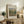 May include: Framed art print featuring Renoir's impressionist landscape painting of lush green chestnut trees in soft brushstrokes, displayed on a rustic wooden sideboard alongside fern plant, stacked books, and ceramic vase in a warm, neutral-toned living room setting with beige upholstered chair visible.