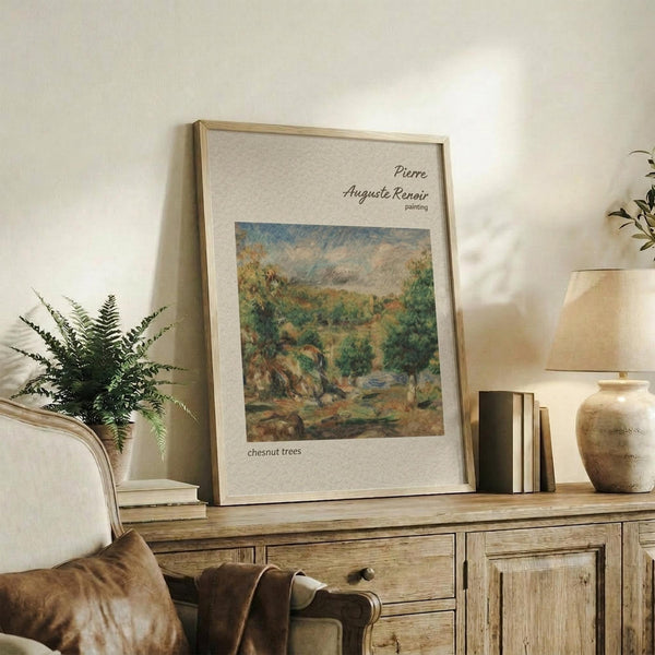 May include: Framed art print featuring Renoir's impressionist landscape painting of lush green chestnut trees in soft brushstrokes, displayed on a rustic wooden sideboard alongside fern plant, stacked books, and ceramic vase in a warm, neutral-toned living room setting with beige upholstered chair visible.