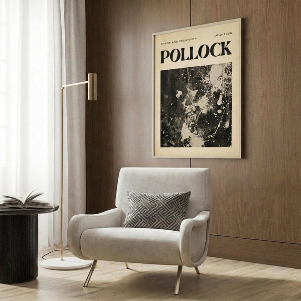 May include: Beige framed art print featuring Jackson Pollock's black and white abstract splatter painting with "POLLOCK" and "CHAOS AND CREATIVITY" text, displayed on wood paneled wall above cream upholstered armchair with geometric patterned pillow, beside brass floor lamp and dark round side table with open book