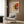 May include: Abstract expressionist poster with bold red, yellow, and black paint splatters and dynamic brushstrokes displayed in cream matting and light wood frame. Hanging on wood-paneled wall above modern gray upholstered armchair with geometric patterned pillow. Contemporary room setting features brass floor lamp, dark round side table with open book, and natural lighting from window with curtains.