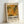 May include: Framed abstract art exhibition poster leaning against white wall. Features Jackson Pollock-style paint splatters in autumn colors - orange, yellow, green, and brown with white drip lines. Text reads "POLLOCK" in bold black letters with "EMBRACE THE CHAOTIC REALM OF CREATIVITY" and dates "MAY 18, 2030 - AUGUST 14, 2030" below. Light wood frame with white matting, soft natural lighting creating gentle shadows.