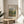 May include: Framed art print displaying Pierre Auguste Renoir's impressionist landscape painting of chestnut trees in autumn colors, featuring green foliage and earthy tones. Print sits on round wooden side table next to beige upholstered chair with plant leaves visible, natural sunlight casting shadows on neutral wall background.