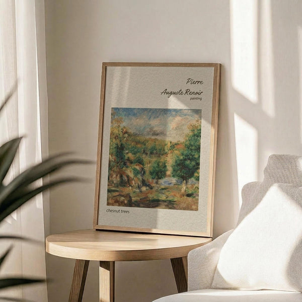 May include: Framed art print displaying Pierre Auguste Renoir's impressionist landscape painting of chestnut trees in autumn colors, featuring green foliage and earthy tones. Print sits on round wooden side table next to beige upholstered chair with plant leaves visible, natural sunlight casting shadows on neutral wall background.
