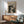 May include: Black and white abstract art poster with paint splatters and drips in wooden frame, hanging above mid-century modern wooden sideboard with white ceramic lamp. Sage green upholstered chair with wooden arms sits nearby on wooden floor. Natural sunlight creates geometric shadows on white wall in minimalist living room setting.