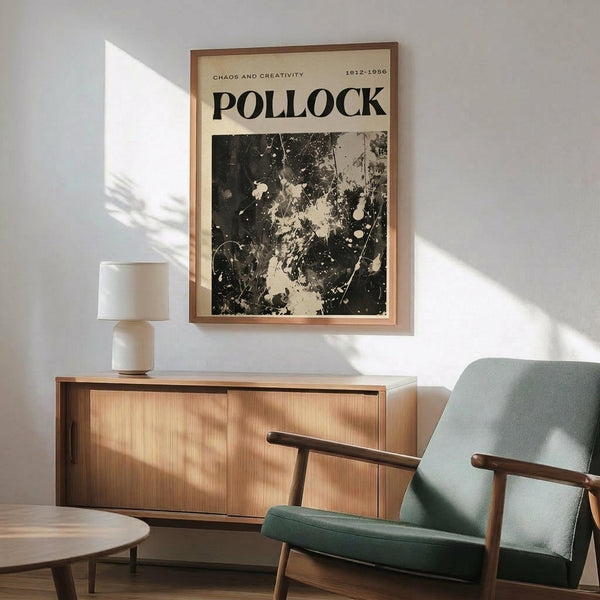 May include: Black and white abstract art poster with paint splatters and drips in wooden frame, hanging above mid-century modern wooden sideboard with white ceramic lamp. Sage green upholstered chair with wooden arms sits nearby on wooden floor. Natural sunlight creates geometric shadows on white wall in minimalist living room setting.