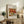 May include: Framed art print featuring Renoir's colorful still life painting of apples, oranges and lemons displayed on rustic wooden sideboard next to fern plant, books, ceramic vase and beige upholstered chair in neutral-toned living room setting