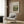 May include: Beige upholstered armchair with geometric patterned throw pillow positioned next to framed Renoir landscape print on wood paneled wall. Brass floor lamp with white circular base stands beside chair. Dark round side table with open book visible on left. Warm neutral tones throughout modern living room setting with natural lighting from window.