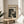 May include: Framed poster with cream background featuring "POLLOCK" in bold black letters, "CHAOS AND CREATIVITY 1912-1956" text, and black and white abstract paint splatter artwork below. Thin wooden frame sits on round wooden side table next to beige upholstered chair, with plant leaves visible in soft natural lighting.