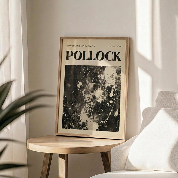 May include: Framed poster with cream background featuring "POLLOCK" in bold black letters, "CHAOS AND CREATIVITY 1912-1956" text, and black and white abstract paint splatter artwork below. Thin wooden frame sits on round wooden side table next to beige upholstered chair, with plant leaves visible in soft natural lighting.