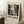 May include: Framed vintage-style exhibition poster leaning against white wall beside cozy chair. Black and white abstract splatter artwork dominates the design with bold "POLLOCK" text and exhibition dates from 2030. Warm lighting creates soft shadows, coffee cup sits on wooden block nearby, cream textured rug below.