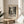 May include: Black and white abstract expressionist poster with paint splatters and drips displayed in thin gold frame on round wooden side table. Cream colored wall backdrop with soft natural lighting creating shadows. Beige upholstered chair partially visible on right side, plant leaves in corner. Exhibition-style poster features bold typography and chaotic paint texture artwork reminiscent of mid-century abstract painting style.