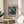 May include: Abstract art poster with swirling teal, purple, and cream marbled patterns in wooden frame, displayed on round wooden stool beside beige upholstered chair. Natural sunlight streams across white walls creating geometric shadows. Green plant leaves visible in corner of minimalist modern interior setting.