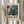 May include: Person holding framed abstract art poster featuring swirling marbled pattern in teal, purple, and cream colors. Exhibition-style design with "POLLOCK" text and dates "MAY 18, 2030 - AUGUST 14, 2030". Light wood frame against neutral background with dried pampas grass visible to the left. Hands wearing cream knit sleeves display the artwork.