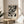 May include: Black and white abstract expressionist artwork with chaotic paint splatters and drips displayed in a light wood frame on a round wooden side table. The poster sits beside a beige upholstered chair with a throw pillow, next to a potted plant with long leaves. Soft natural light creates geometric shadows on cream-colored walls in this minimalist modern living room setting.