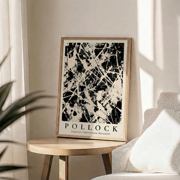 May include: Black and white abstract expressionist artwork with chaotic paint splatters and drips displayed in a light wood frame on a round wooden side table. The poster sits beside a beige upholstered chair with a throw pillow, next to a potted plant with long leaves. Soft natural light creates geometric shadows on cream-colored walls in this minimalist modern living room setting.