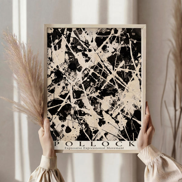 May include: Person in cream ruffle-sleeve top holding framed abstract expressionist artwork featuring black paint splatters and drips on beige background, standing near dried pampas grass against soft neutral curtained window setting
