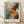 May include: A framed abstract art print with dynamic paint splatters and brushstrokes in vibrant orange, red, turquoise, black and cream colors, displayed against a white textured wall on light wood flooring. The wooden frame sits on the floor with dramatic shadows cast across the wall, showcasing the energetic expressionist style artwork with bold color contrasts and fluid paint movement.