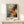 May include: Abstract expressionist artwork in wooden frame leaning against white wall on light wood console table. The painting features dynamic splashes and drips in orange, red, black, white and blue-gray tones creating energetic movement across the canvas. A small white wrapped gift with evergreen sprig sits in front of the framed piece. Soft natural lighting creates gentle shadows on the minimalist white wall backdrop.