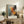 May include: Abstract expressionist art print in light wood frame displaying energetic paint splatters and brushstrokes in orange, red, black, and blue tones against cream background. Poster sits on rustic wooden sideboard next to fern plant, ceramic vase, and stacked books in warmly lit living room with beige armchair.