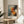 May include: Abstract expressionist artwork poster in wooden frame, featuring dynamic splashes and drips in orange, red, black, and blue against cream background, displayed on round wooden side table next to beige upholstered chair with plant leaves visible, warm natural lighting creating soft shadows on neutral wall.