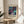 May include: Abstract art poster with swirling purple, teal and blue cosmic design featuring golden dots and flowing lines, displayed in light wood frame on round wooden side table next to beige upholstered chair. Soft natural lighting creates shadows on cream wall with white curtains and green plant leaves visible in bright, minimalist living room setting.