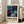 May include: Framed abstract art poster leaning against white wall next to beige sofa in bright living room. Artwork features swirling cosmic design with deep purples, teals, and navy blues, scattered with golden circles and flowing lines. White matted frame displays exhibition-style poster with bold black text below the celestial abstract image. Natural sunlight streams through tall windows onto wooden floors.
