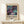 May include: Framed poster with abstract cosmic artwork featuring swirling purple, teal, and dark blue colors with golden dots and flowing lines, displayed on light wood shelf against cream wall. Small wrapped gift box with feather sits in front of natural wood frame.
