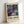 May include: Framed abstract art poster with swirling purple, teal and dark blue cosmic patterns, gold circular dots and flowing lines throughout. Features exhibition-style text layout with "POLLOCK" in bold letters and "EMBRACE THE CHAOTIC REALM OF CREATIVITY" subtitle, plus dates "MAY 18, 2030 - AUGUST 14, 2030". Light wooden frame sits propped against white wall on white surface, casting soft shadow.
