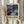 May include: Person holding framed abstract art poster featuring swirling cosmic design in deep purples, teals, and blues with gold splatter accents. White wooden frame against neutral curtained background with dried wheat stems visible on left side.